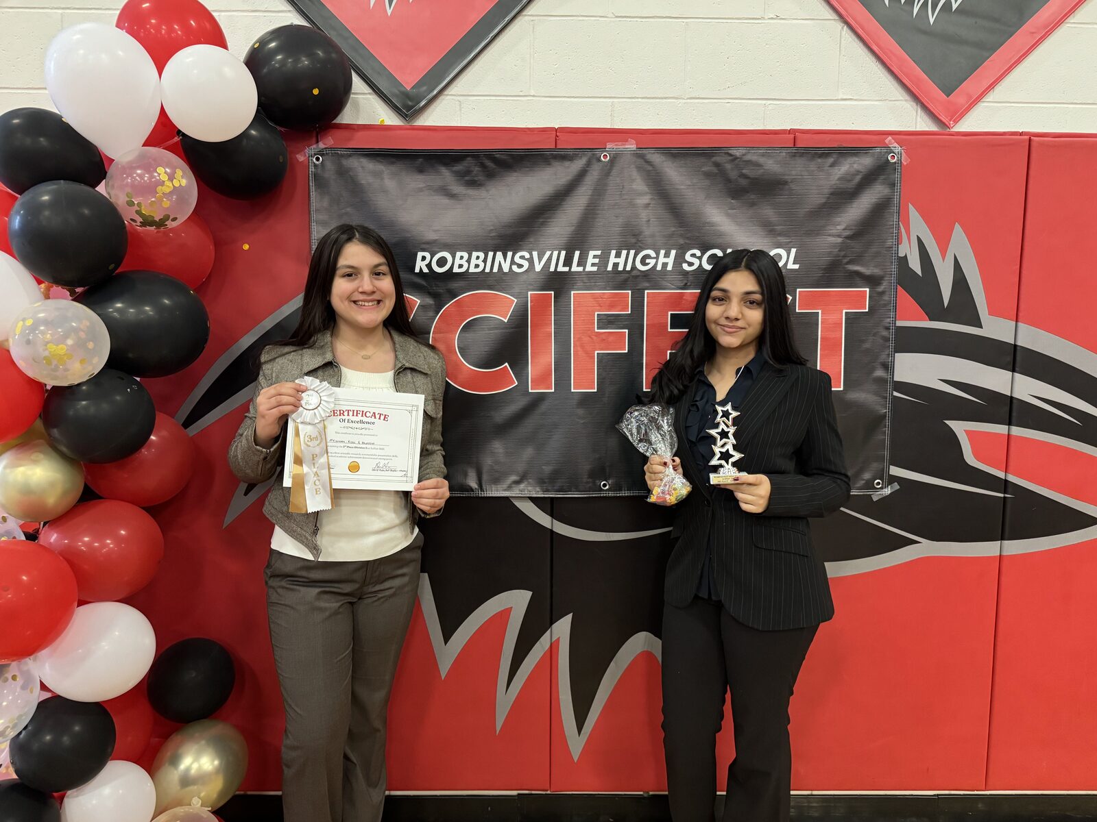 Yana Chauhan and Alis Onat hold their 3rd Place Division 1 ribbons, certificate, and trophy in front of the SciFest banner.