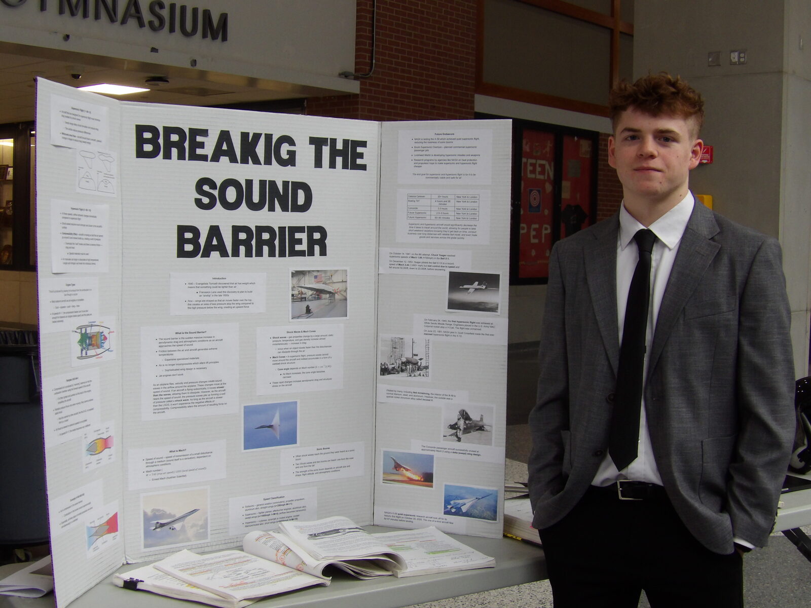 A SciFest 2026 presenter stands beside his tri-fold board, Breaking the Sound Barrier.